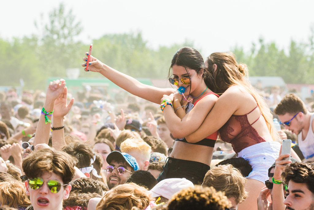 Dos jóvenes disfrutando de un concierto al aire libre entre una multitud, tomando selfies y abrazándose