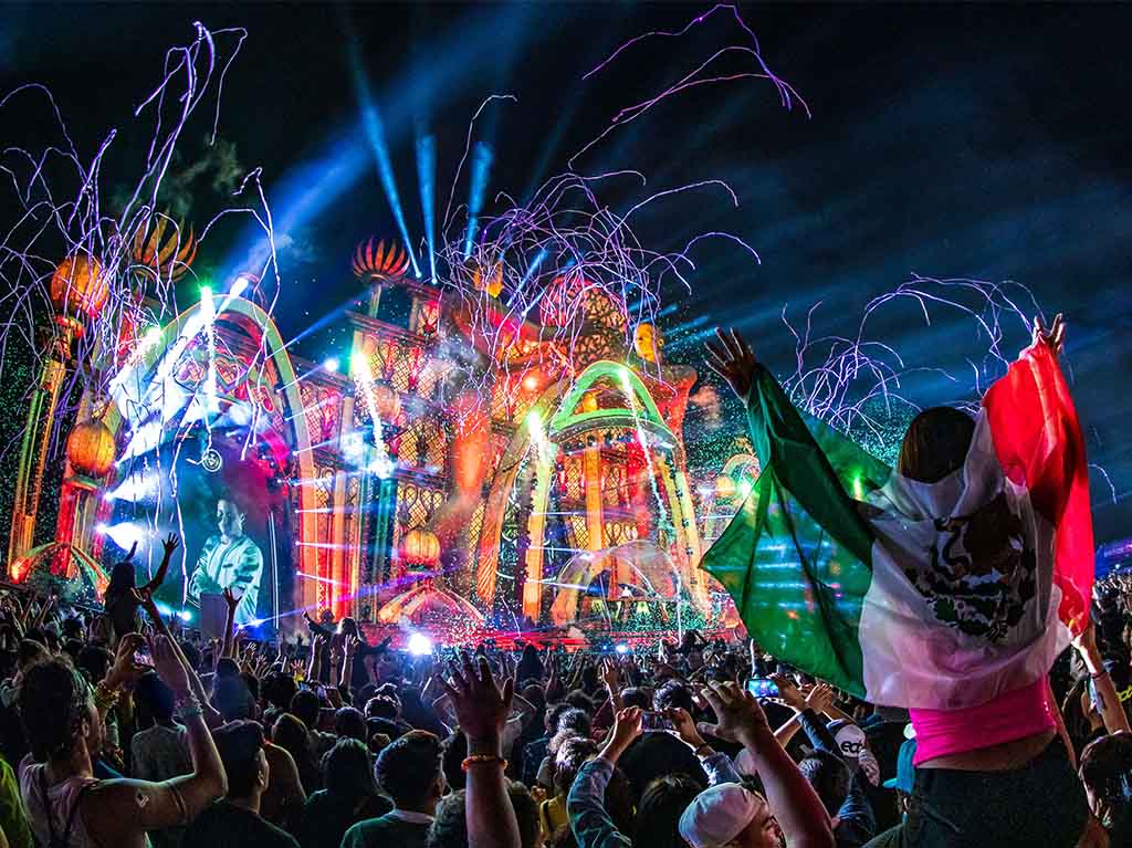 Multitud disfrutando de un festival nocturno con luces vibrantes, efectos visuales y una persona ondeando una bandera de México