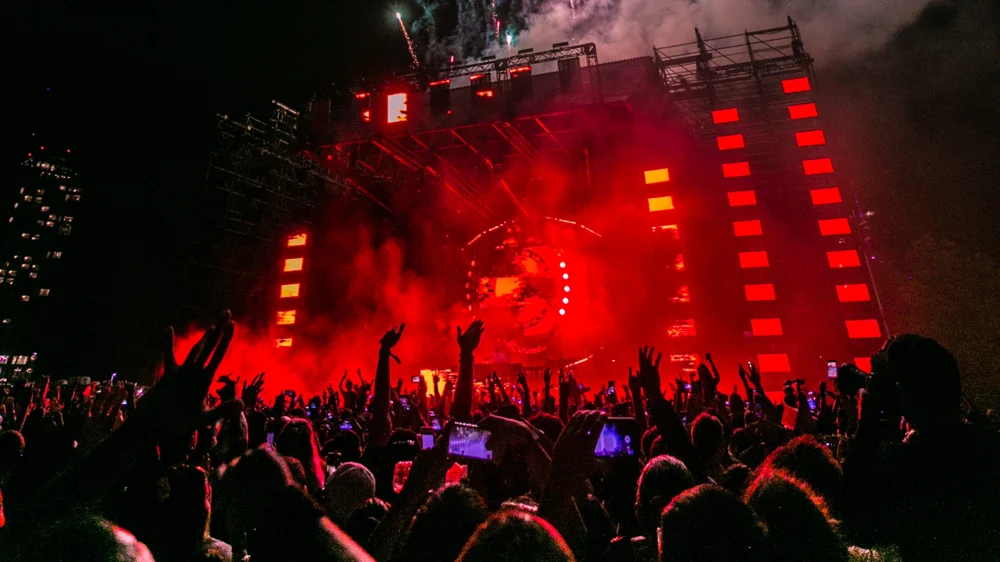 Escenario iluminado con luces rojas en un concierto nocturno, rodeado de una multitud animada con manos levantadas
